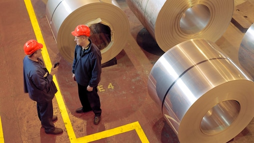 Two factory workers standing next to rolls of steel
