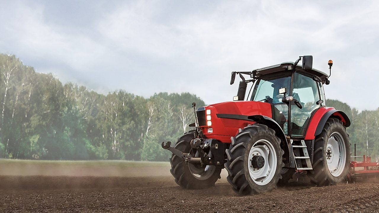 A red tractor ploughing a muddy field with trees in the background