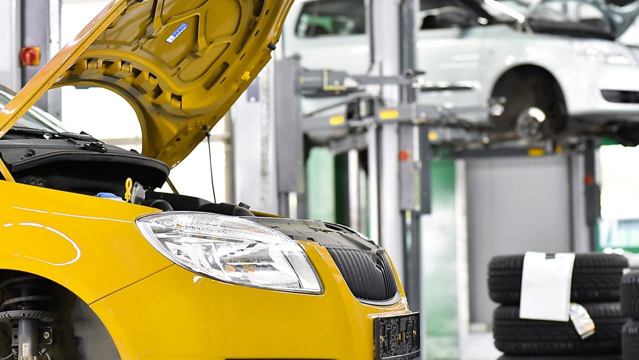 Two cars in a garage, one yellow and one white, with their bonnets open, and some car tyres in the background