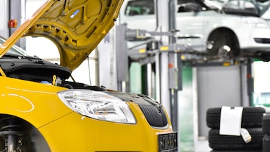 Two cars in a garage, one yellow and one white, with their bonnets open, and some car tyres in the background